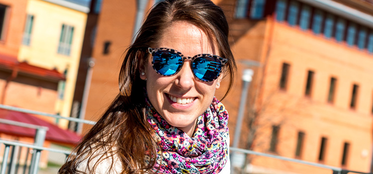 Picture of a girl standing in front of the University of Skövde. She smiles and wears sunglasses.