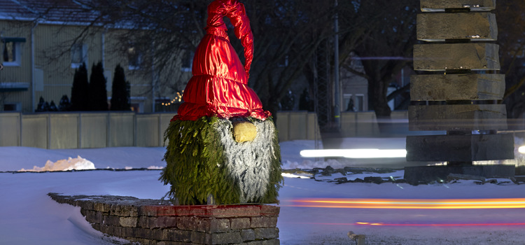 Bild på en tomte som är tillverkad av granris. Tomten står i snön och har en röd luva på huvudet.