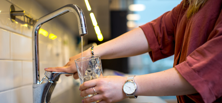 En person tar häller upp vatten i ett dricksglas från en kran. 